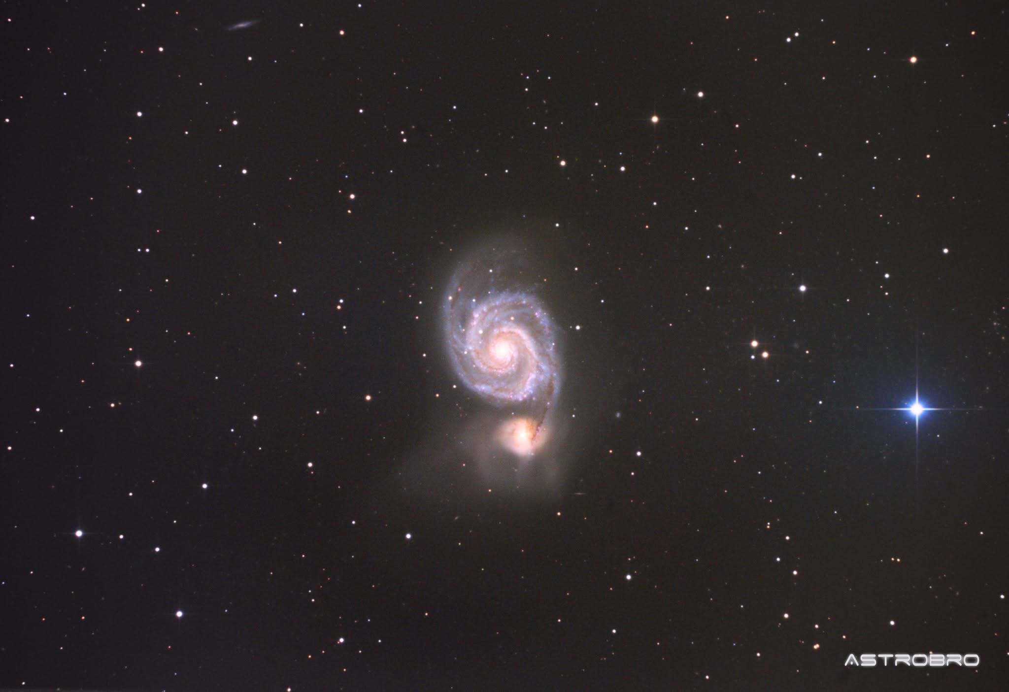 Galaxie du Tourbillon (M51) photographiée au télescope : spirale vue de face aux bras bien dessinés, reliée par un pont de matière à une petite galaxie compagne jaunâtre, dans les Chiens de Chasse.