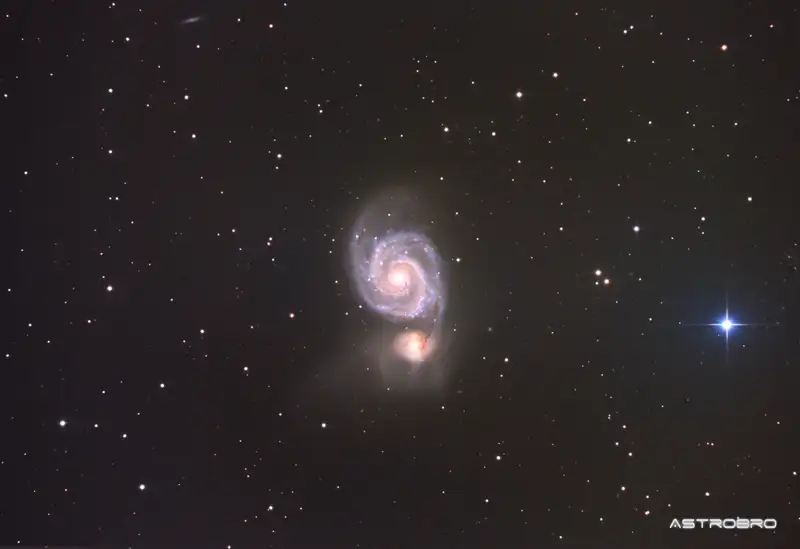 Galaxie du Tourbillon (M51) photographiée au télescope : spirale vue de face aux bras bien dessinés, reliée par un pont de matière à une petite galaxie compagne jaunâtre, dans les Chiens de Chasse.