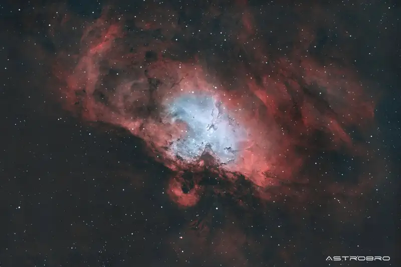 Nébuleuse de l'Aigle (M16) photographiée au télescope : nuage d'hydrogène rouge étendu entourant une cavité bleutée où se détachent les Piliers de la Création, colonnes de poussière sombre.