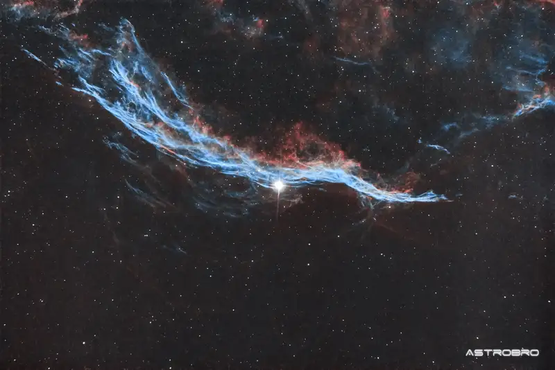 Nébuleuse du Balai de Sorcière (NGC 6960) photographiée au télescope : long ruban de filaments rouges et bleus traversant le champ de part en part, longeant une étoile brillante, dans le Cygne.