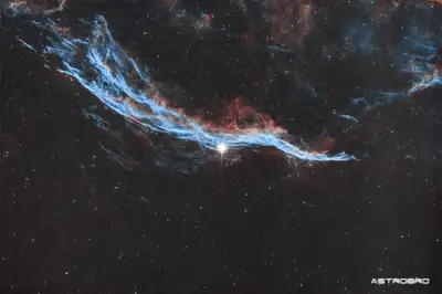 Nébuleuse du Balai de Sorcière (NGC 6960) photographiée au télescope : long ruban de filaments rouges et bleus traversant le champ de part en part, longeant une étoile brillante, dans le Cygne.
