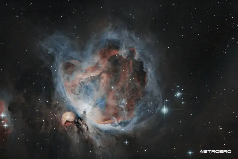 Nébuleuse d'Orion (M42) photographiée au télescope : vaste nuage de gaz bleu et rose entourant l'amas du Trapèze, avec la nébuleuse de l'Homme qui court en bas à gauche, sur fond d'étoiles.