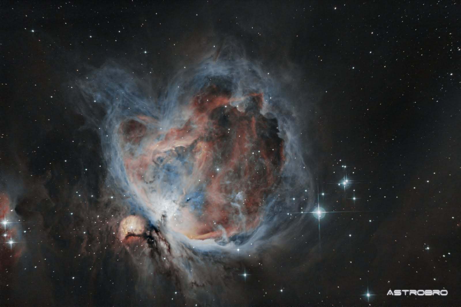 Nébuleuse d'Orion (M42) photographiée au télescope : vaste nuage de gaz bleu et rose entourant l'amas du Trapèze, avec la nébuleuse de l'Homme qui court en bas à gauche, sur fond d'étoiles.
