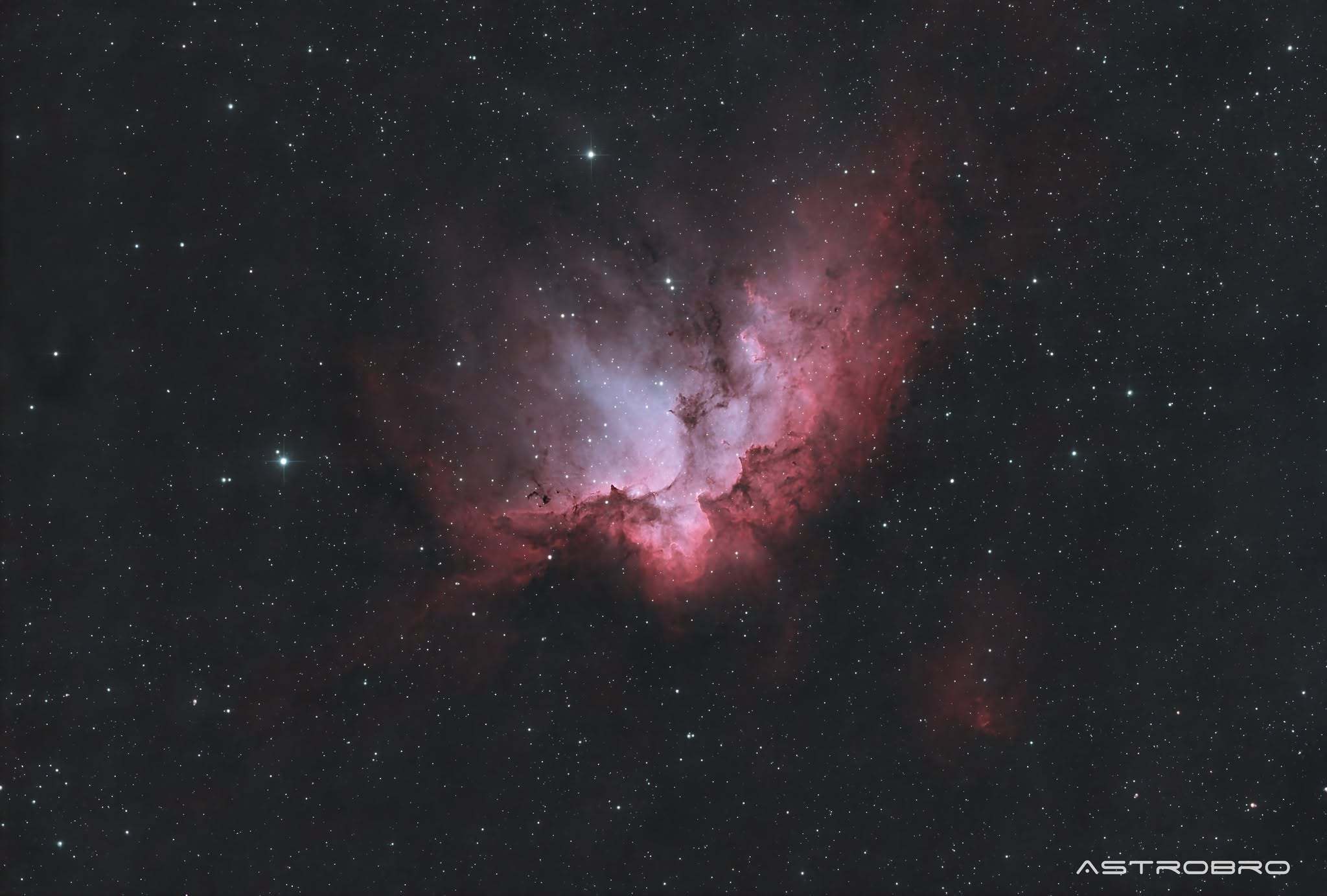 Nébuleuse du Sorcier (NGC 7380) photographiée au télescope : nuage d'hydrogène rouge et rose dessinant une silhouette encapuchonnée, avec un amas d'étoiles jeunes au centre et des bandes de poussière sombre.